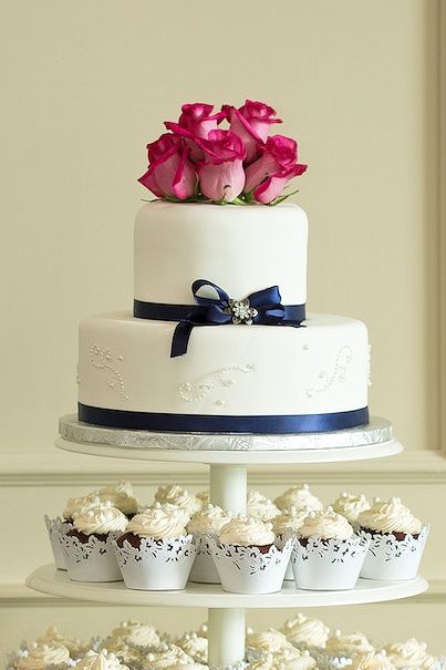 A two tier wedding cake. The lwoer tier is bigger and the bottom of each tier is wrapped with a one-inch thick ribbon. The cake stands on a cake display structure, with cupcakes around it.