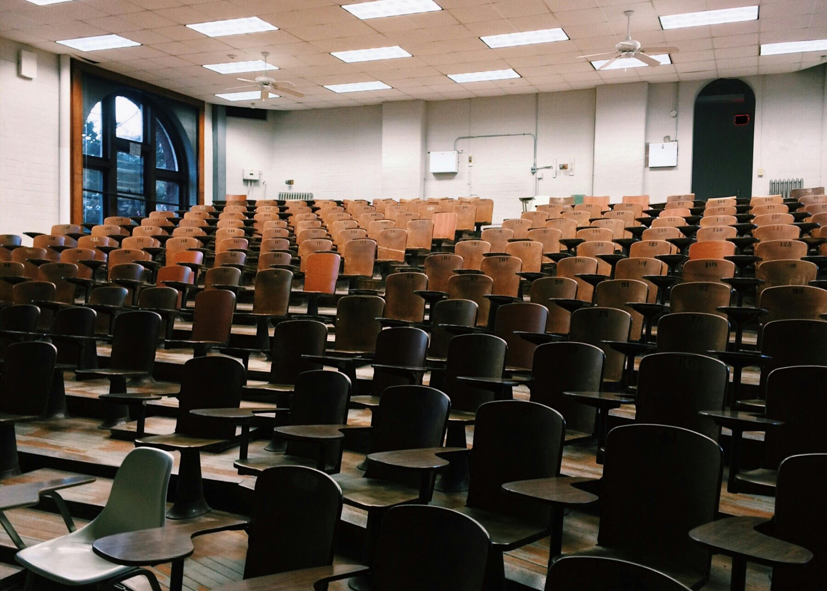 An empty, university-style auditorium, with tiers of single seats and small writing desks, facing up towards the back of the room CC0
